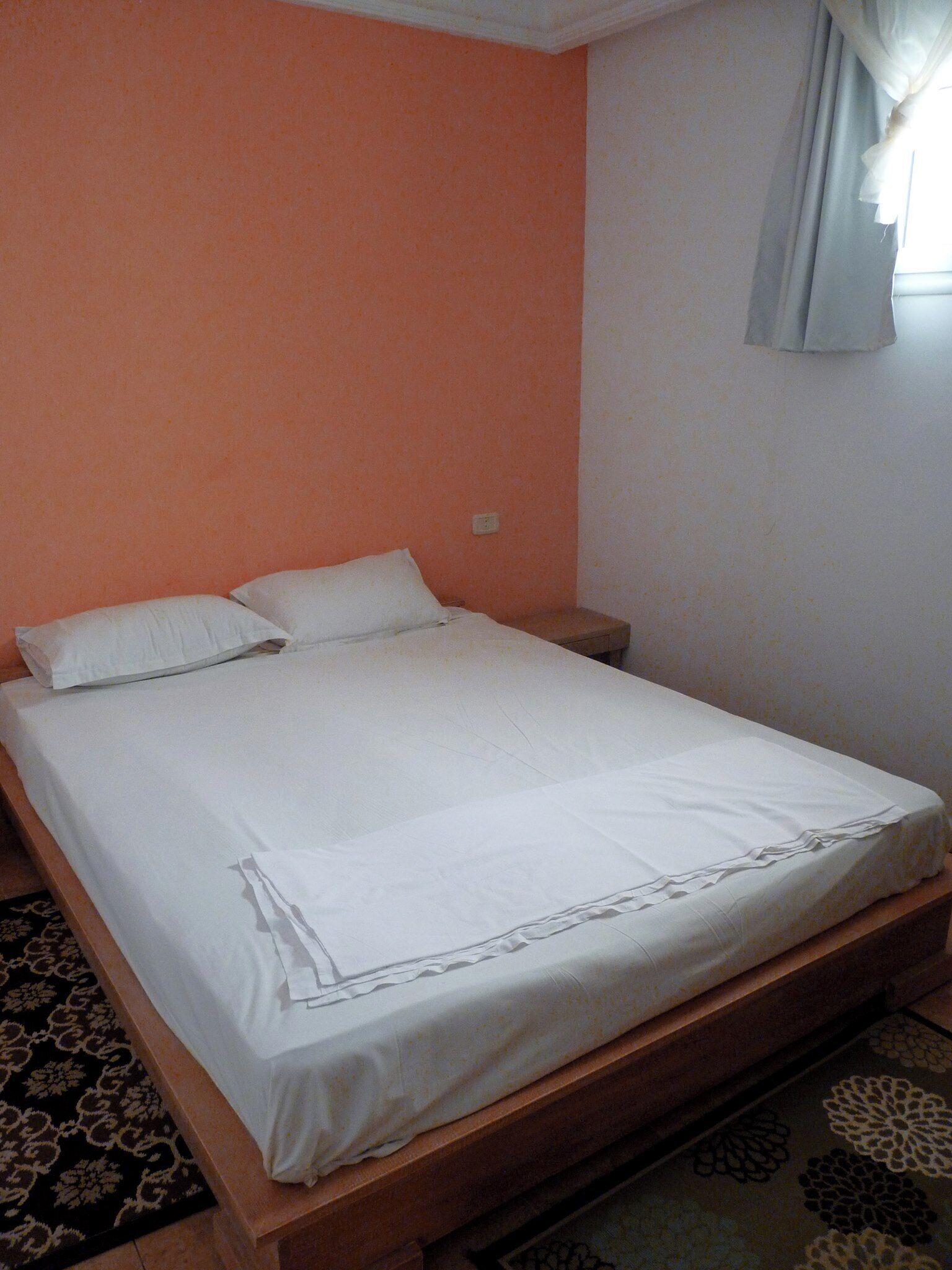Un lit simple dans une chambre dont un mur est orange, avec une couvre-lit blanche et un tapis à motifs.