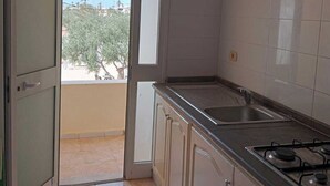 A kitchen with a gas stove, a sink, and cabinets. There is a door leading to a balcony with a view of trees and buildings. - Résidence Le Phare (Djerba Midun)