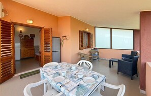 Ein Essbereich mit Tisch für vier Personen, eine Küche mit Kühlschrank und ein Fenster mit Jalousien.