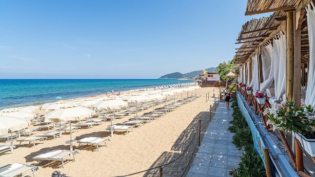 Ein Strand mit Liegestühlen und Sonnenschirmen, einem wolkenlosen, blauen Himmel und einem Gebäude mit Bambusdach.