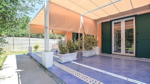 Ein überdachter Patio-Bereich mit Kübelpflanzen, Fliesenboden und einer grünen Tür.