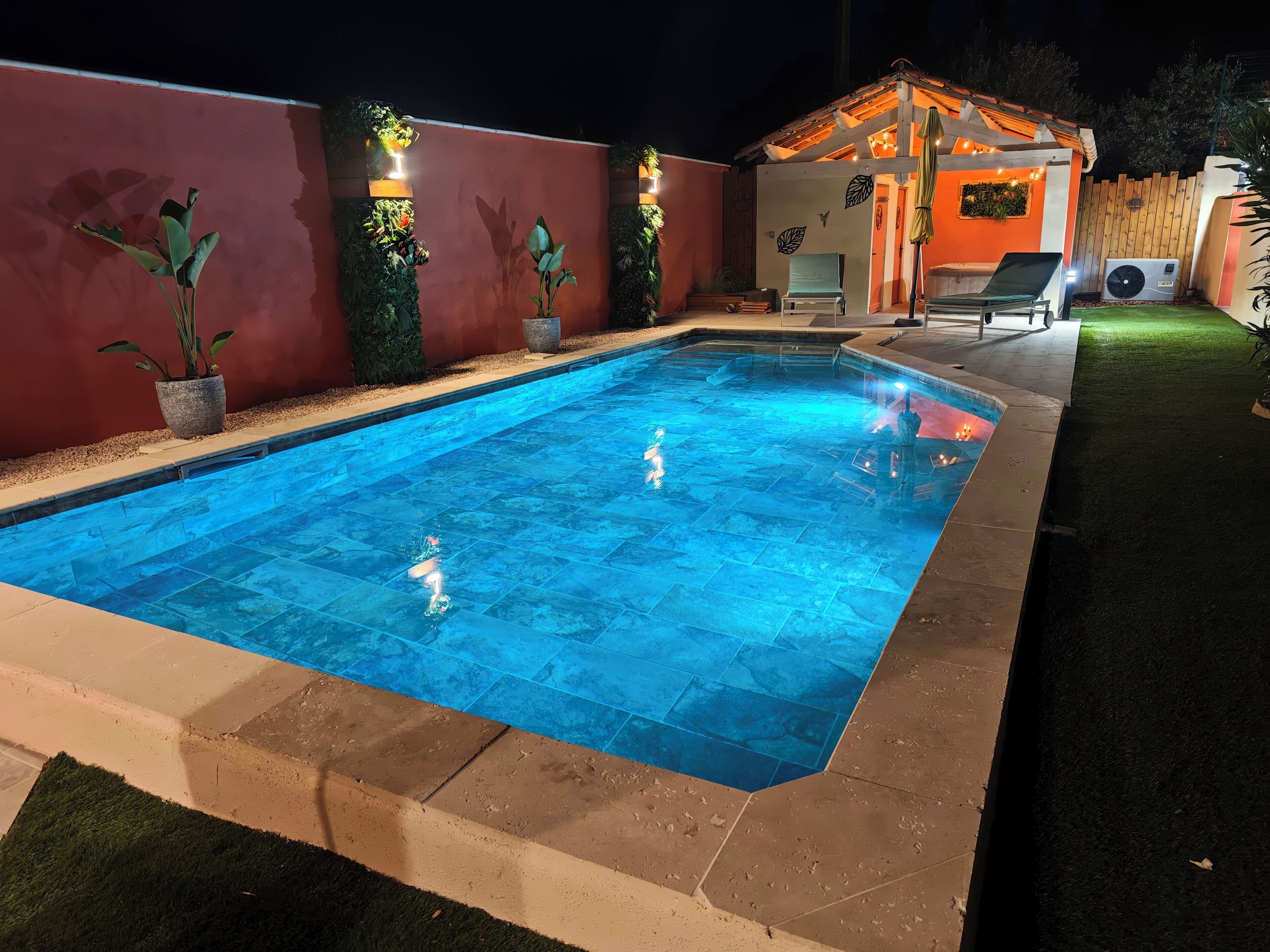 A swimming pool with a tiled deck, surrounded by a red wall and a wooden pergola with seating.