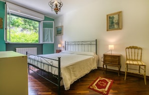 Ein Schlafzimmer mit Bett, Stuhl, Nachttisch, einem Fenster mit Vorhängen, einer Heizung und einem Bild an der Wand.