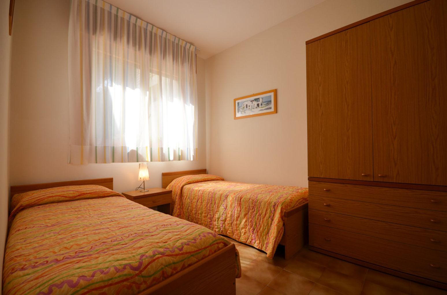 Un dormitorio con dos camas, una mesita de noche con lámpara, un armario de madera y una ventana con cortinas translúcidas.