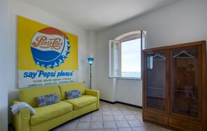 Une pièce avec un canapé jaune, un meuble en bois, une grande affiche publicitaire pour Pepsi Cola et une fenêtre donnant sur un paysage.