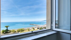 Blick aus einem Fenster auf eine Küstenlandschaft mit blauem Himmel, einem steinernen Wellenbrecher und einer grünen Fläche mit Pflanzen.