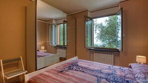Un dormitorio con un espejo grande, una cama con colcha estampada, una silla y una ventana con vistas a árboles.