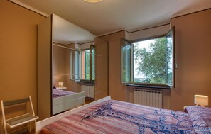 Ein Schlafzimmer mit einem großen Spiegel, einem Bett mit gemustertem Überwurf, einem Sessel und einem Fenster mit Blick auf Bäume.