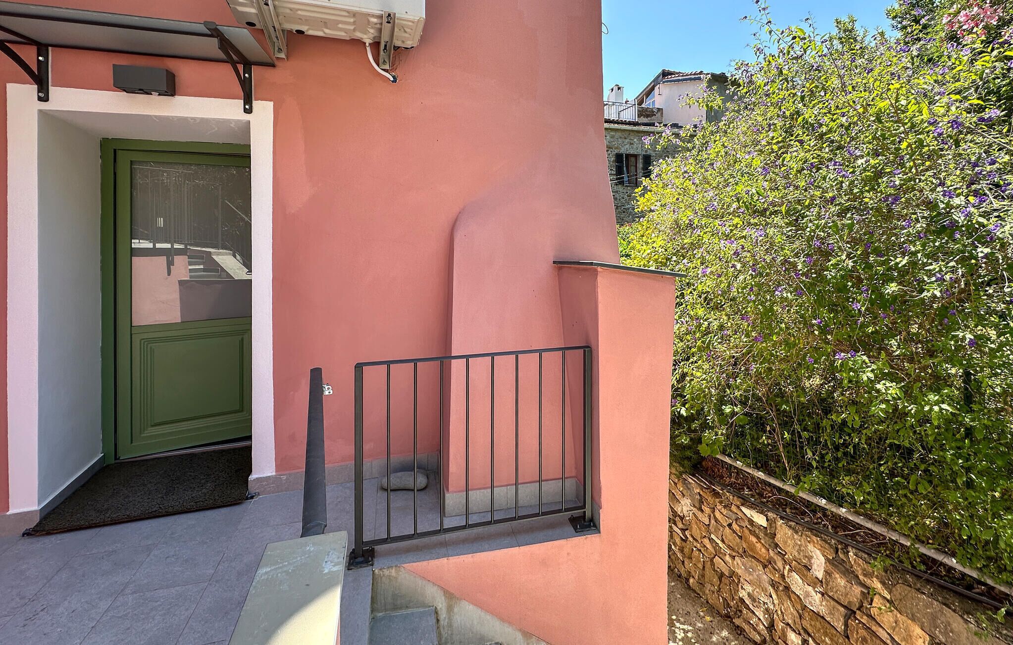 Un bâtiment rose avec une porte verte, un portail en métal et un mur de pierre orné d’un arbuste en fleurs.