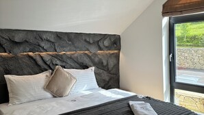 A bedroom with a stone headboard, a bed with white and beige bedding, and a view of greenery through a window.