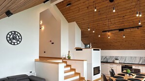 A modern interior with a wooden ceiling, a staircase leading to an upper level, a kitchen, and a seating area with a sofa and tables.