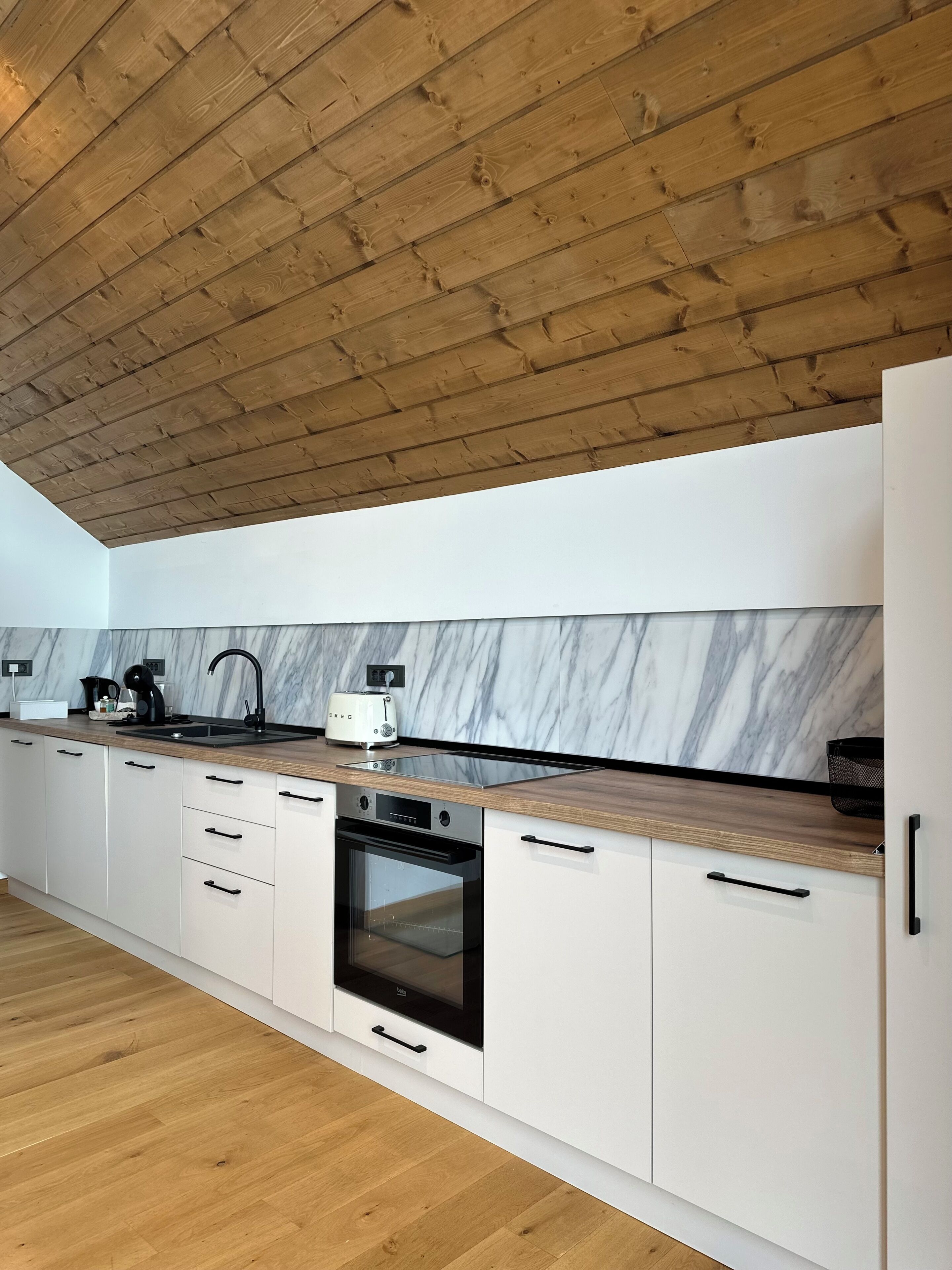 Una cocina moderna con vigas de madera en el techo, muebles blancos, un protector contra salpicaduras de mármol y un horno empotrado.