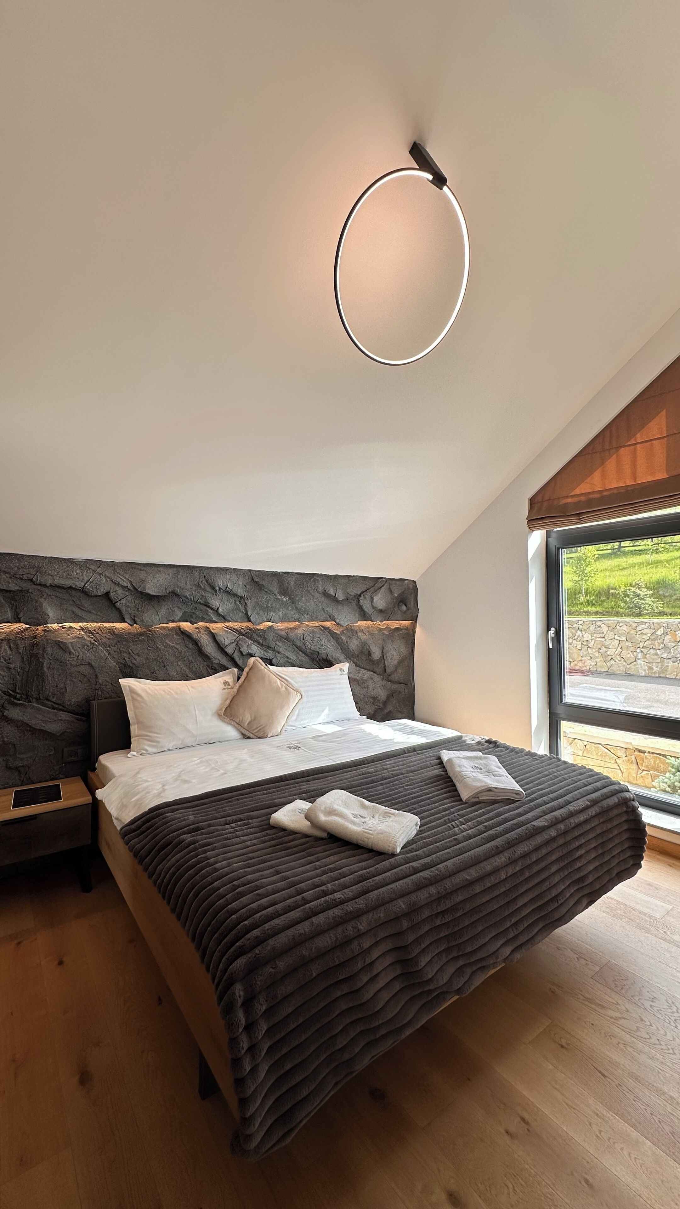 Ein Schlafzimmer mit einem steinernen Kopfende, einem Bett mit dunkler Decke, einem Nachttisch und einem Fenster mit Blick auf Grünflächen.