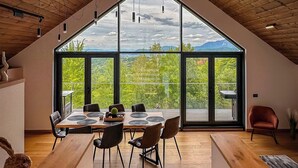 A spacious dining area with a high ceiling, wooden beams, and large windows offering a view of greenery and mountains.