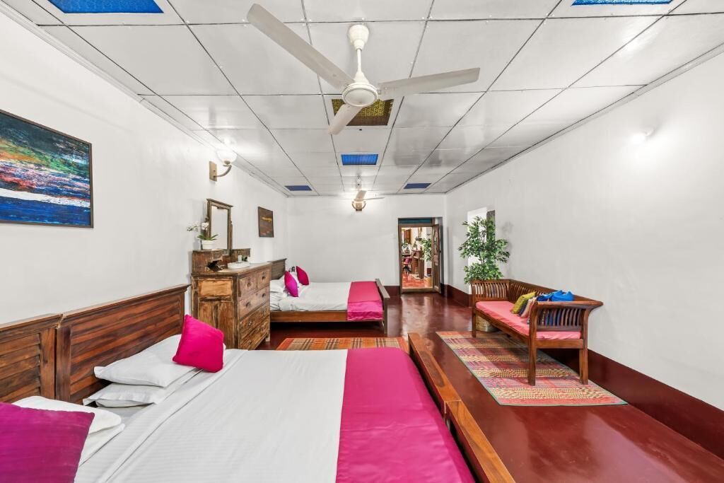 A hotel room with a bed, a wooden bench, a desk, and a ceiling fan.