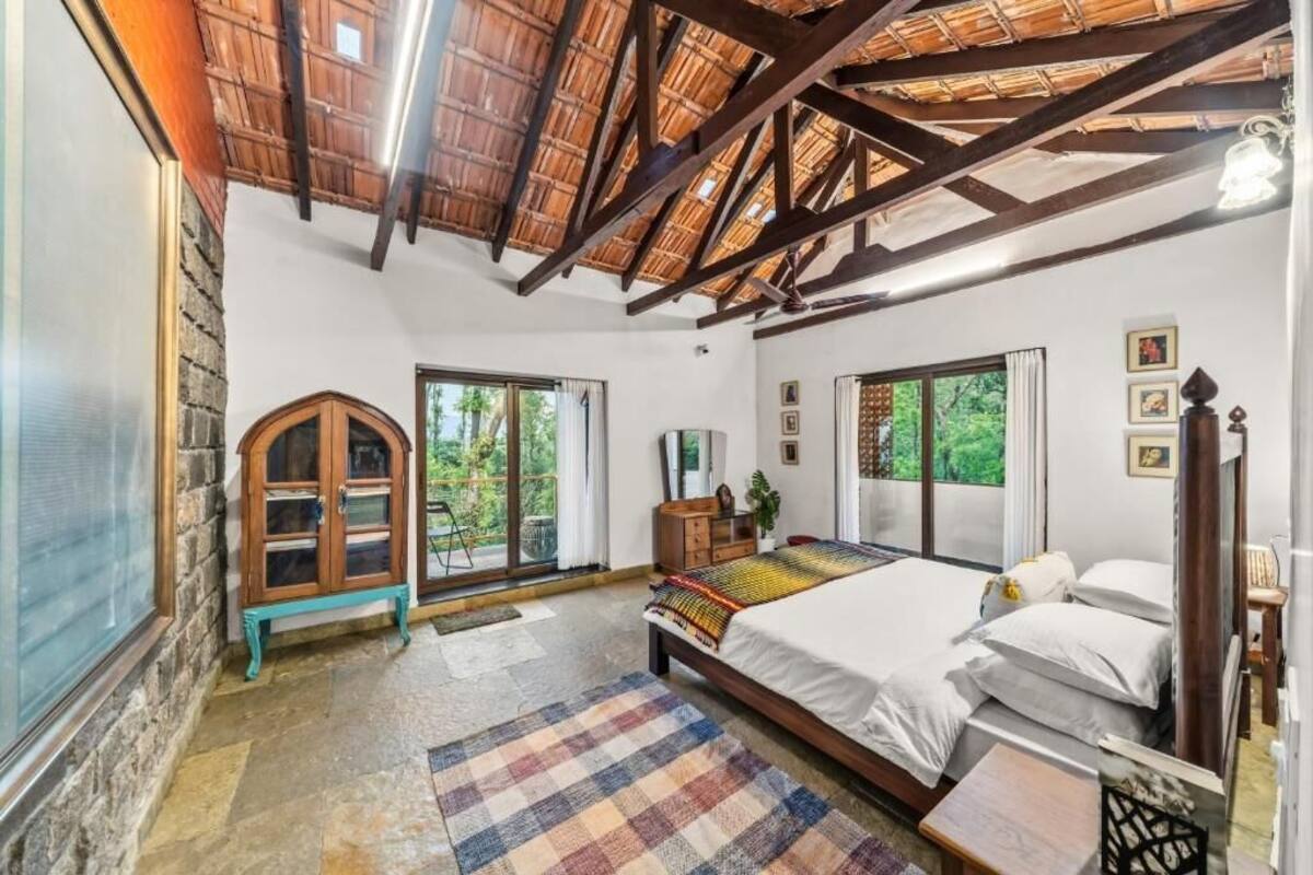 A bedroom with a large bed, wooden beams, a stone wall, and a view of greenery.