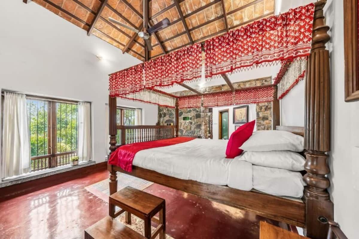 A room with a wooden bed, a bench, a window with curtains, and a stone wall.