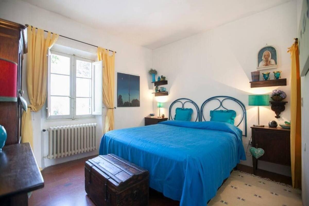 Un dormitorio con cama, un cofre de madera, un radiador, una ventana con cortinas y una estantería de pared con objetos decorativos.