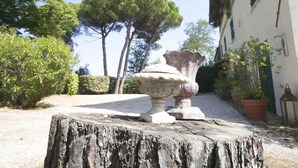 A rustic outdoor setting with a stone urn on a tree stump, surrounded by greenery and a building with green shutters. - Casale novilara (Novilara)