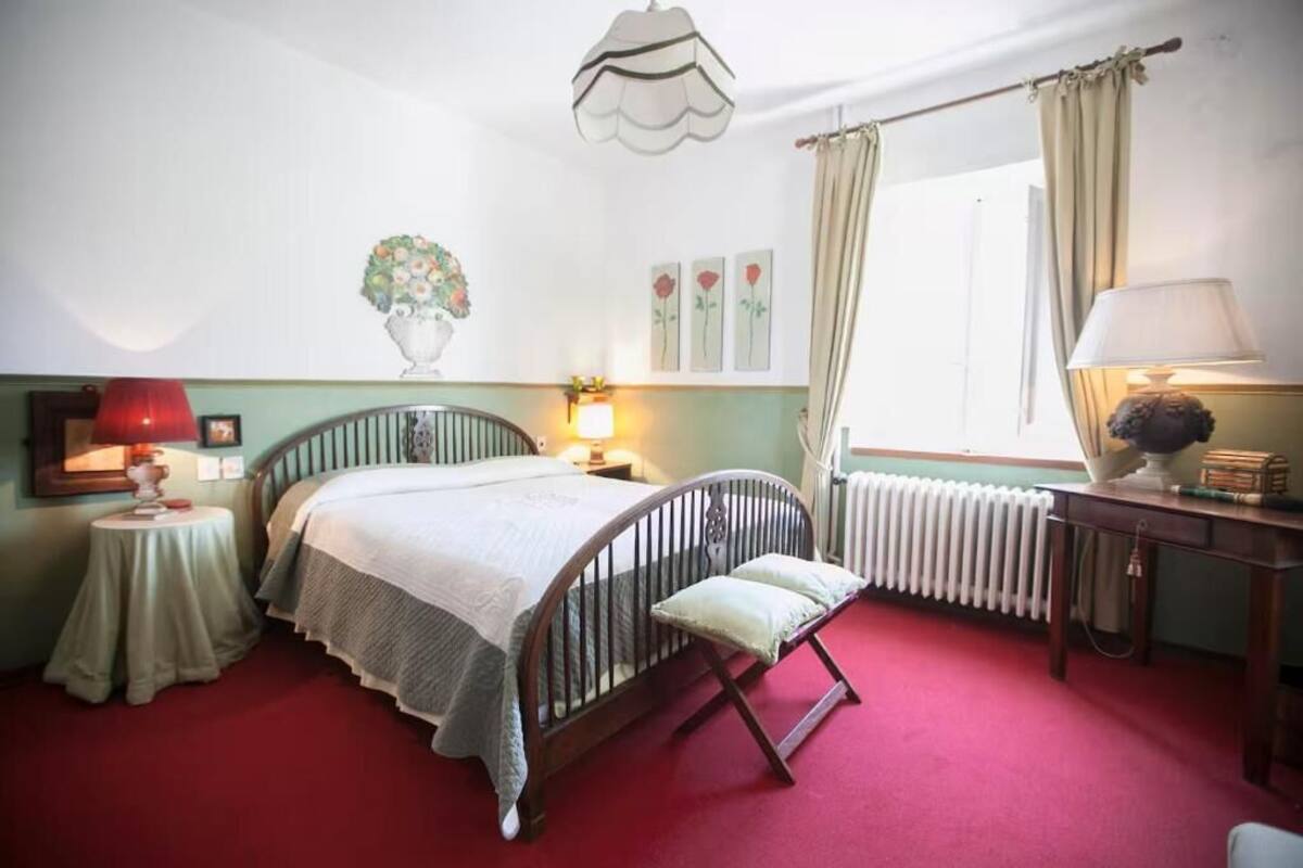 Un dormitorio con cama, mesitas de noche, un radiador, una ventana con cortinas y una lámpara.