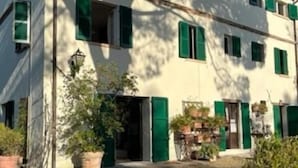 A two-story building with green shutters, a courtyard with potted plants, and a clear blue sky. - Casale novilara (Novilara)