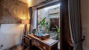 A bathroom with a bathtub, a wooden vanity with a tray of toiletries, a patterned wall, and a view of an outdoor area with plants. - Kasubatu (City)