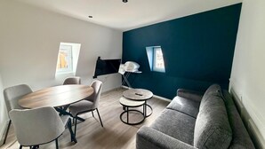 A modern living room with a dining table, chairs, a sofa, a TV, and a small table with a lamp. - Cosy studio - Hypercentre - Renovated - Calm and chic (Thonon-les-Bains)