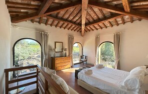 Ein Schlafzimmer mit Holzboden, einem Bett, einem Schminktisch, einem Sessel und zwei Fenstern mit Blick auf Grünflächen.