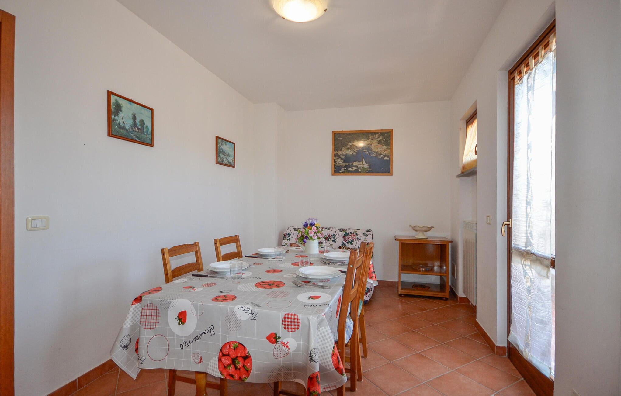 A dining area with a table set for four, wooden chairs, a floral tablecloth, and wall-mounted paintings.