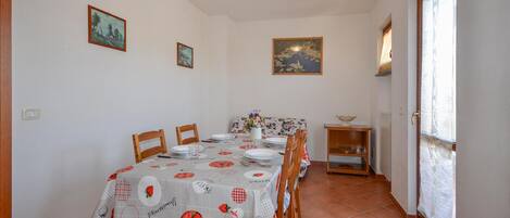 A dining area with a table set for four, wooden chairs, a floral tablecloth, and wall-mounted paintings.