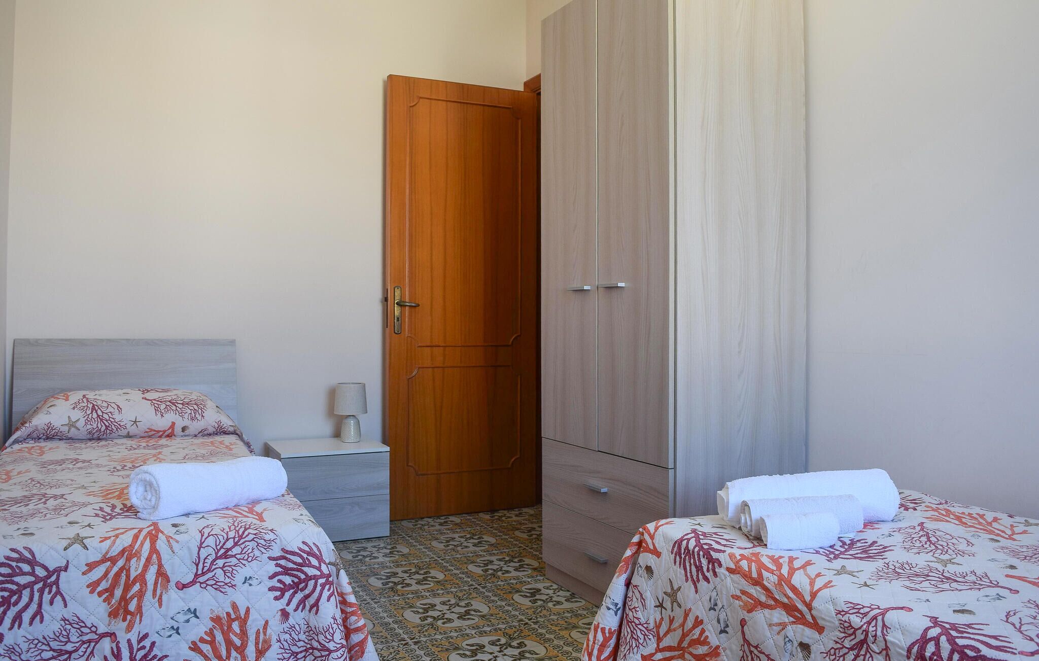 A bedroom with two beds, a wooden wardrobe, a nightstand, and a lamp.