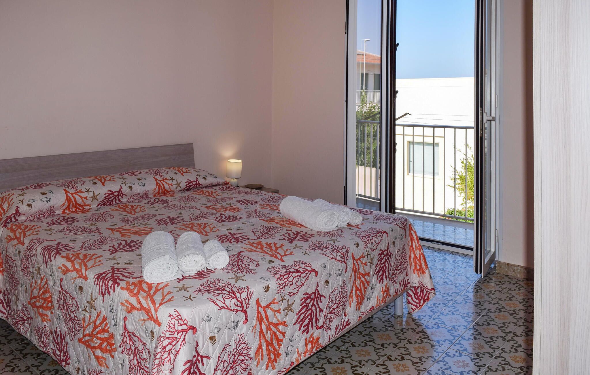A hotel room with a bed, a nightstand, a lamp, and a view of the outside.