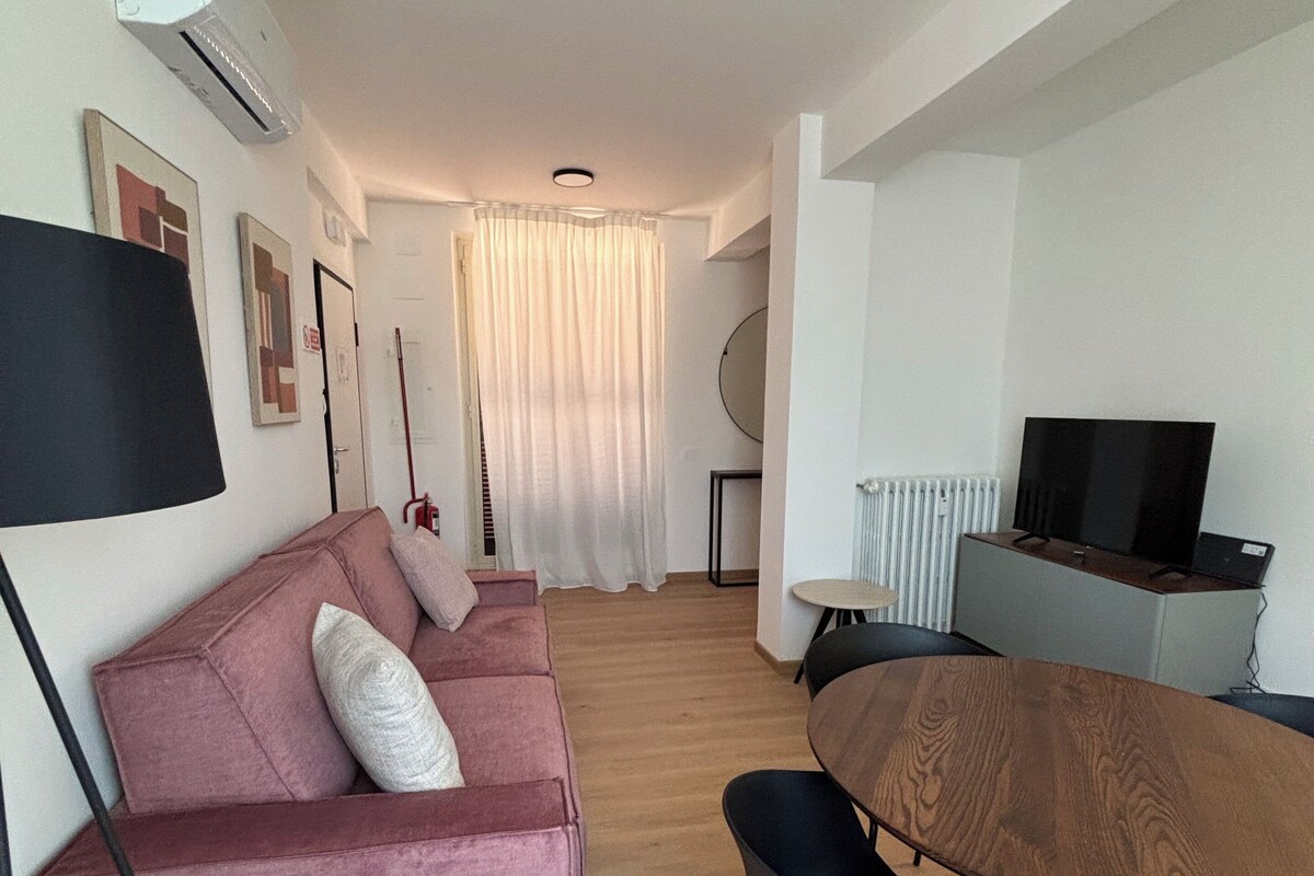 A modern living room with a pink sofa, a wooden dining table, and a TV.