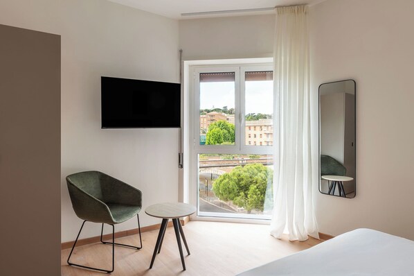 Habitación de hotel con cama, televisor montado en la pared, una silla, una mesita redonda y un ventanal con vistas a edificios y árboles.