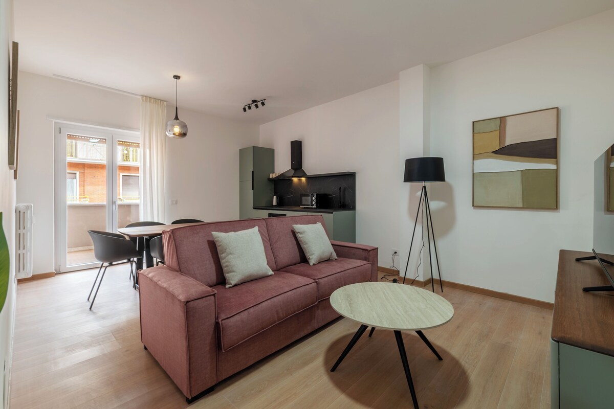 A modern living room with a sofa, a round table, a kitchen area, and a dining set.