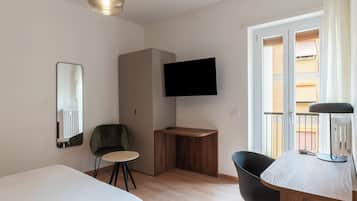 A modern hotel room with a bed, a desk, a chair, a TV, and a window with a view of buildings.