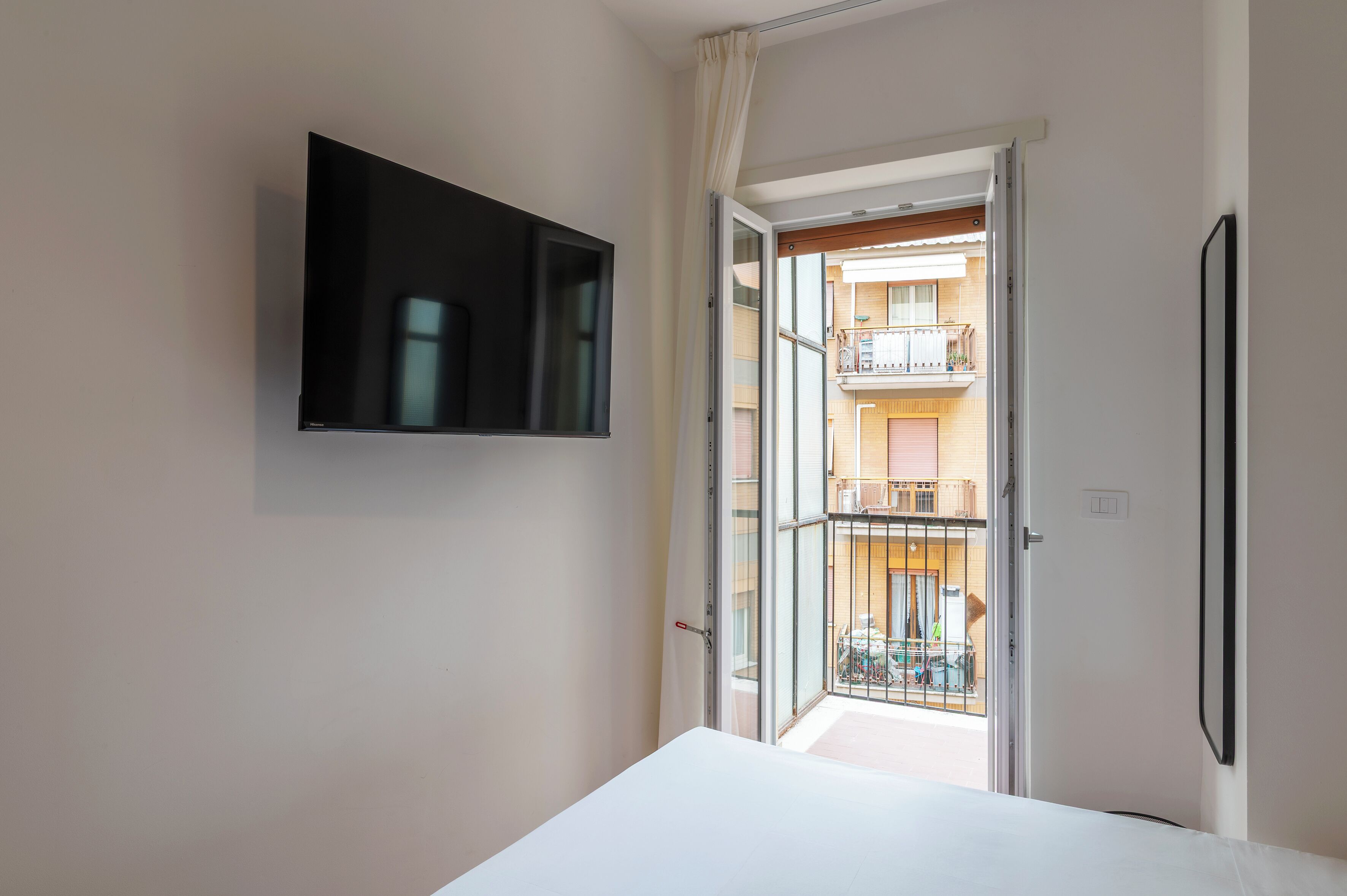 Una stanza con un letto, una TV fissata al muro e una porta aperta su un balcone con vista su un edificio.