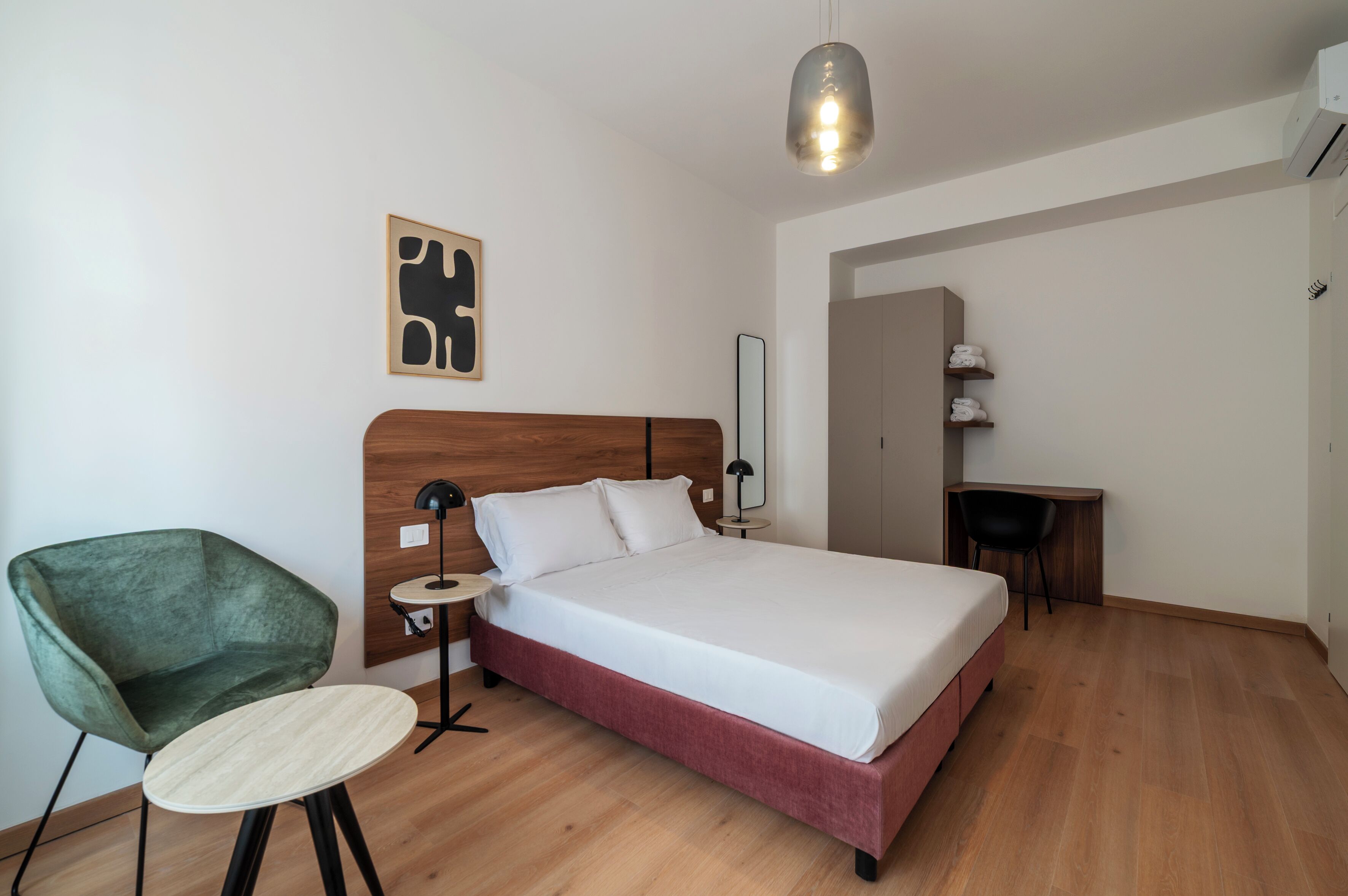 A hotel room with a bed, a green armchair, a small round table, a bedside lamp, and a wall-mounted artwork.