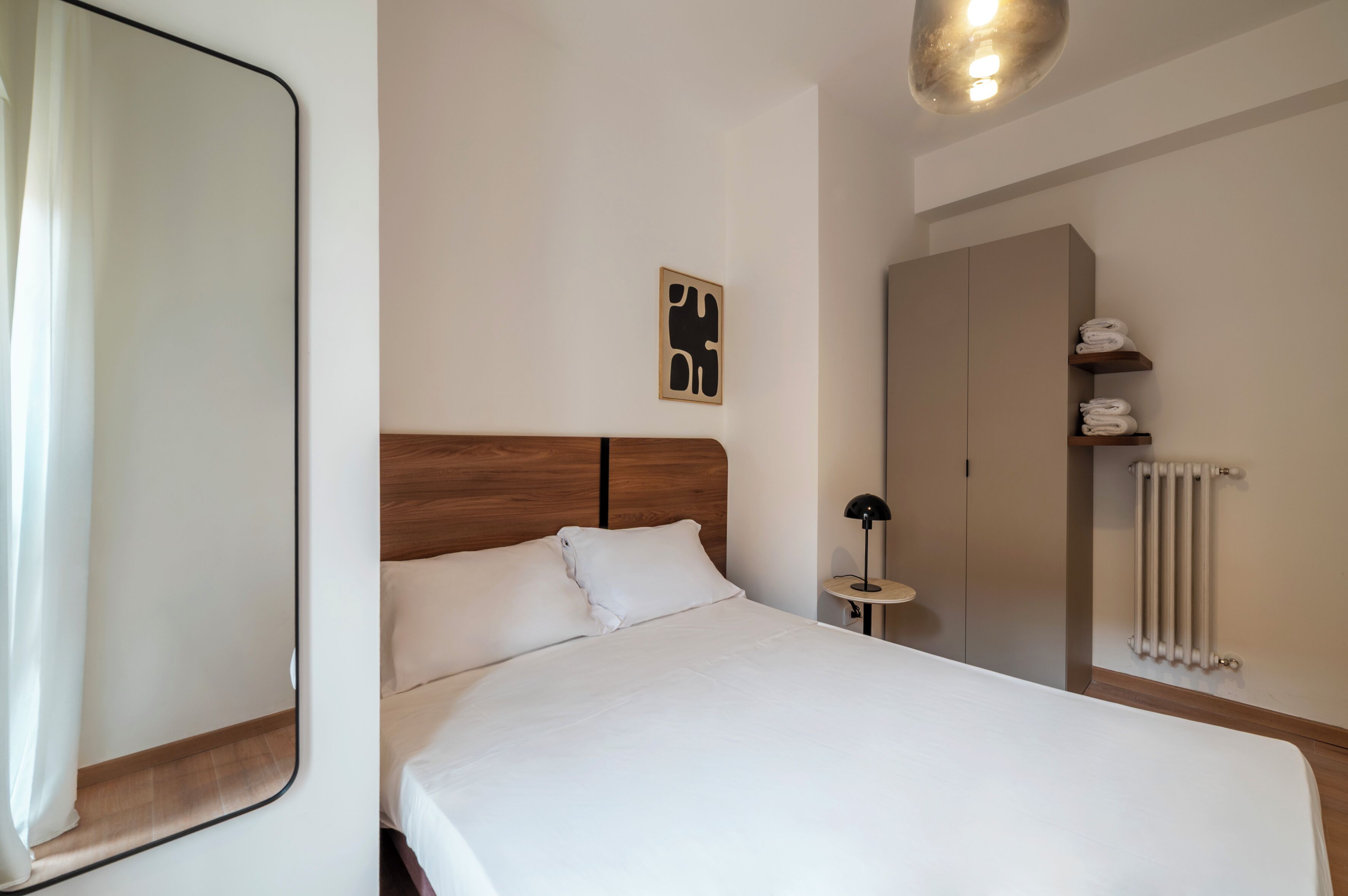 A neatly made bed with white linens, a wooden headboard, a bedside table with a lamp, a radiator, and a wall-mounted artwork.