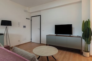 A minimalist living room with a flat-screen TV, a round wooden table, a sofa, a lamp, and a potted plant. - Stylish Balcony Flats by the Vatican (Roma)