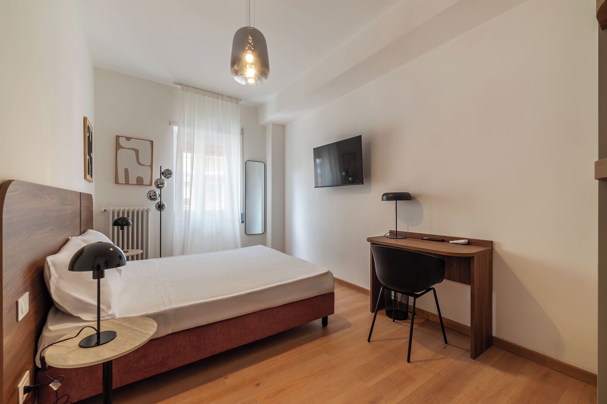 Una camera da letto con un letto, una lampada da comodino, una scrivania, una sedia, una TV e un termosifone.