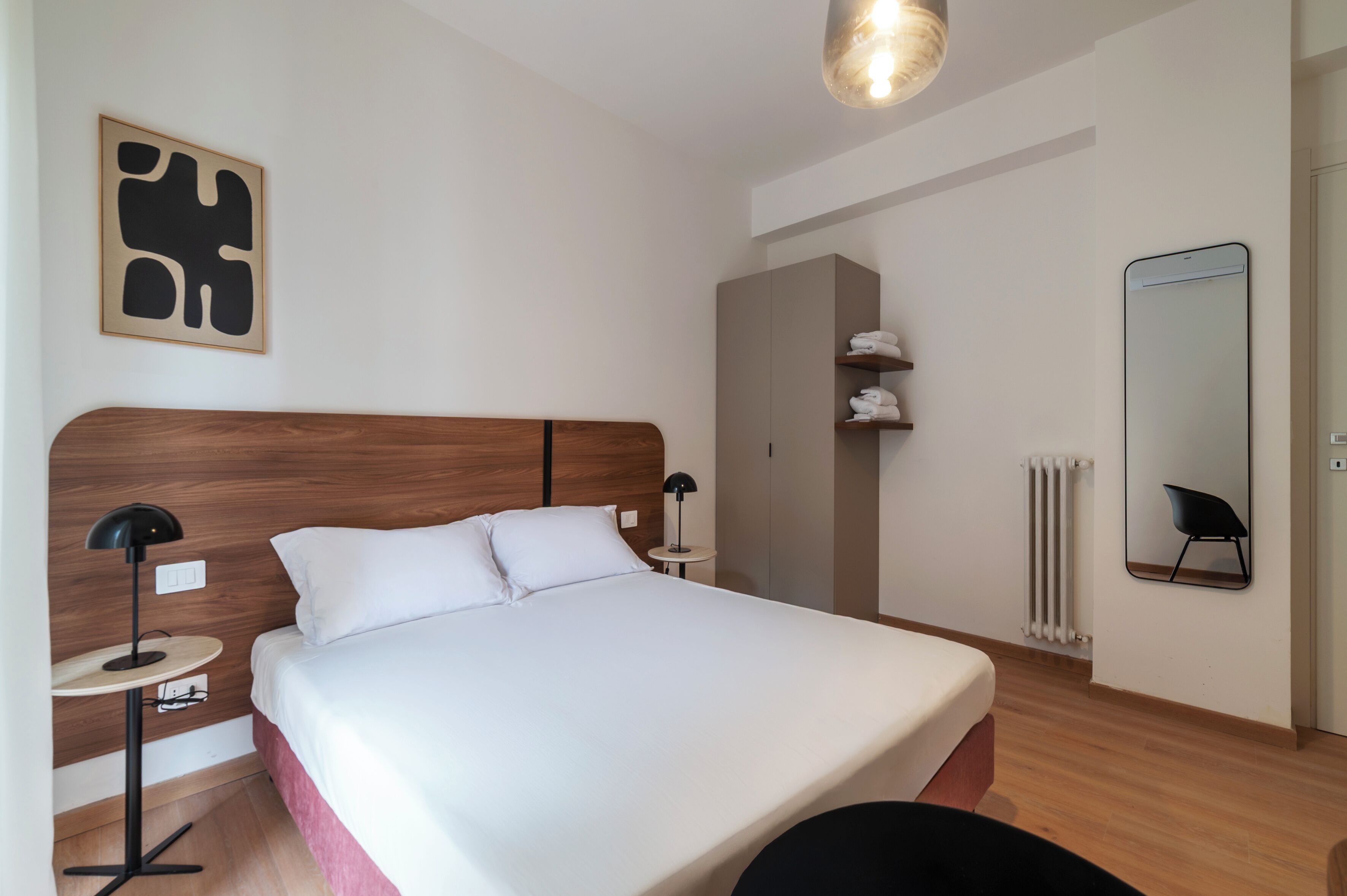 Ein modernes Hotelzimmer mit einem Bett mit Holz-Kopfteil, einem runden Nachttisch mit Lampe, einem Kleiderschrank, einem Heizkörper und einem Spiegel an der Wand.