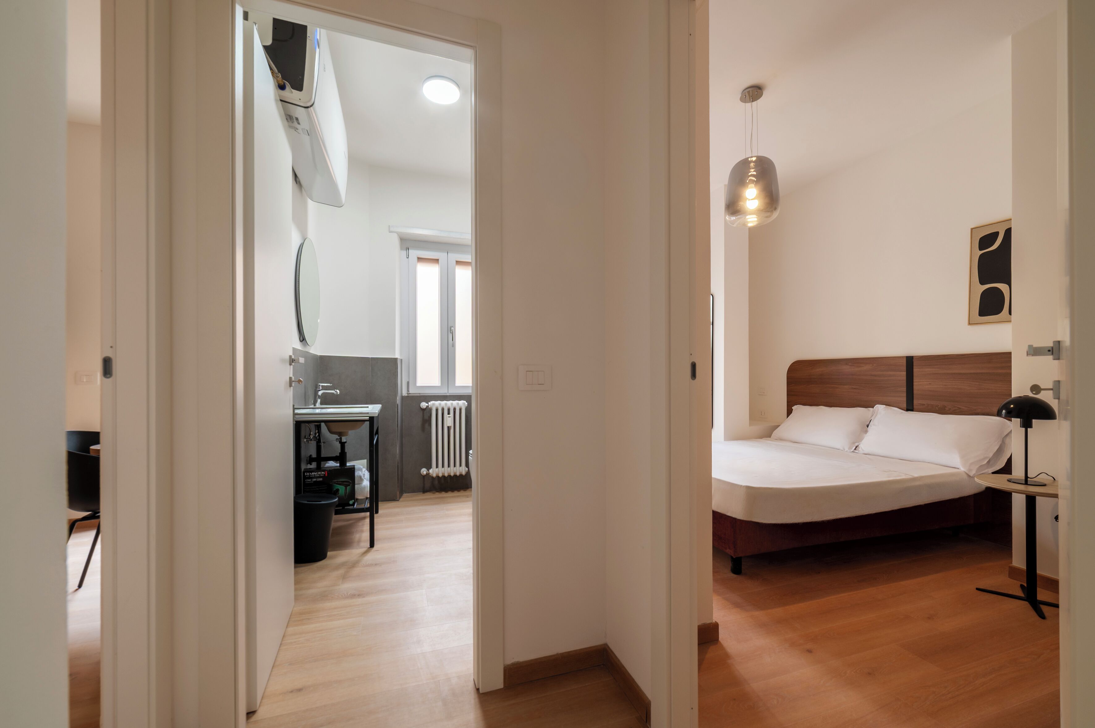 Een slaapkamer met een bed, nachtkastje, lamp en uitzicht op de keuken en het balkon.