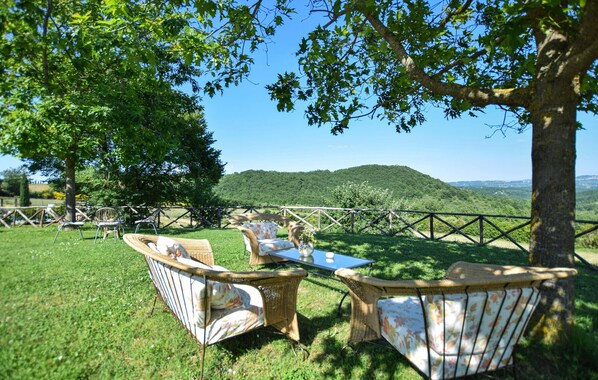 Ein mit Gras belegter Außenbereich mit Korbstühlen und einem Tisch, umgeben von Bäumen und Blick auf eine Hügelkette.