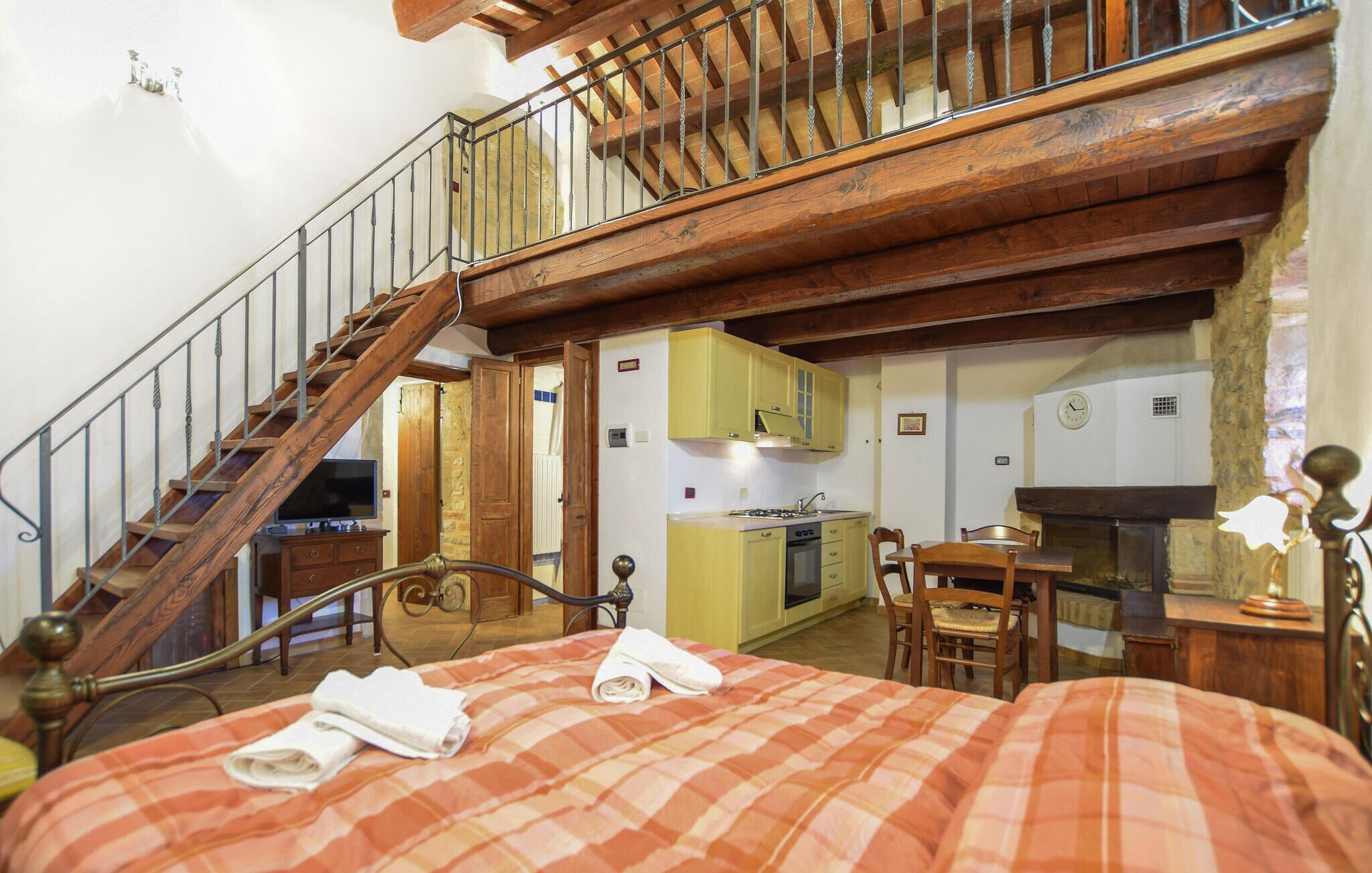 A bedroom with a wooden staircase, a bed with a plaid bedspread, a kitchen area with yellow cabinets, and a dining table with chairs.