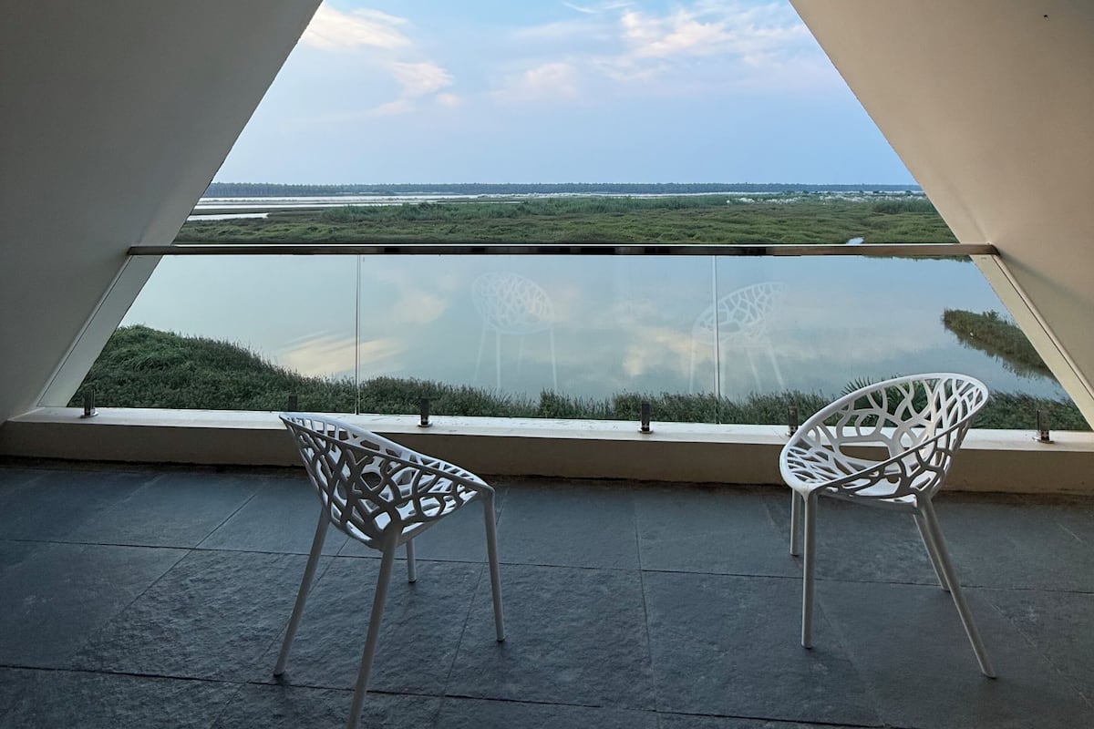 Una habitación con un amplio ventanal triangular que da a un paisaje, dos sillas blancas y suelo de baldosas.