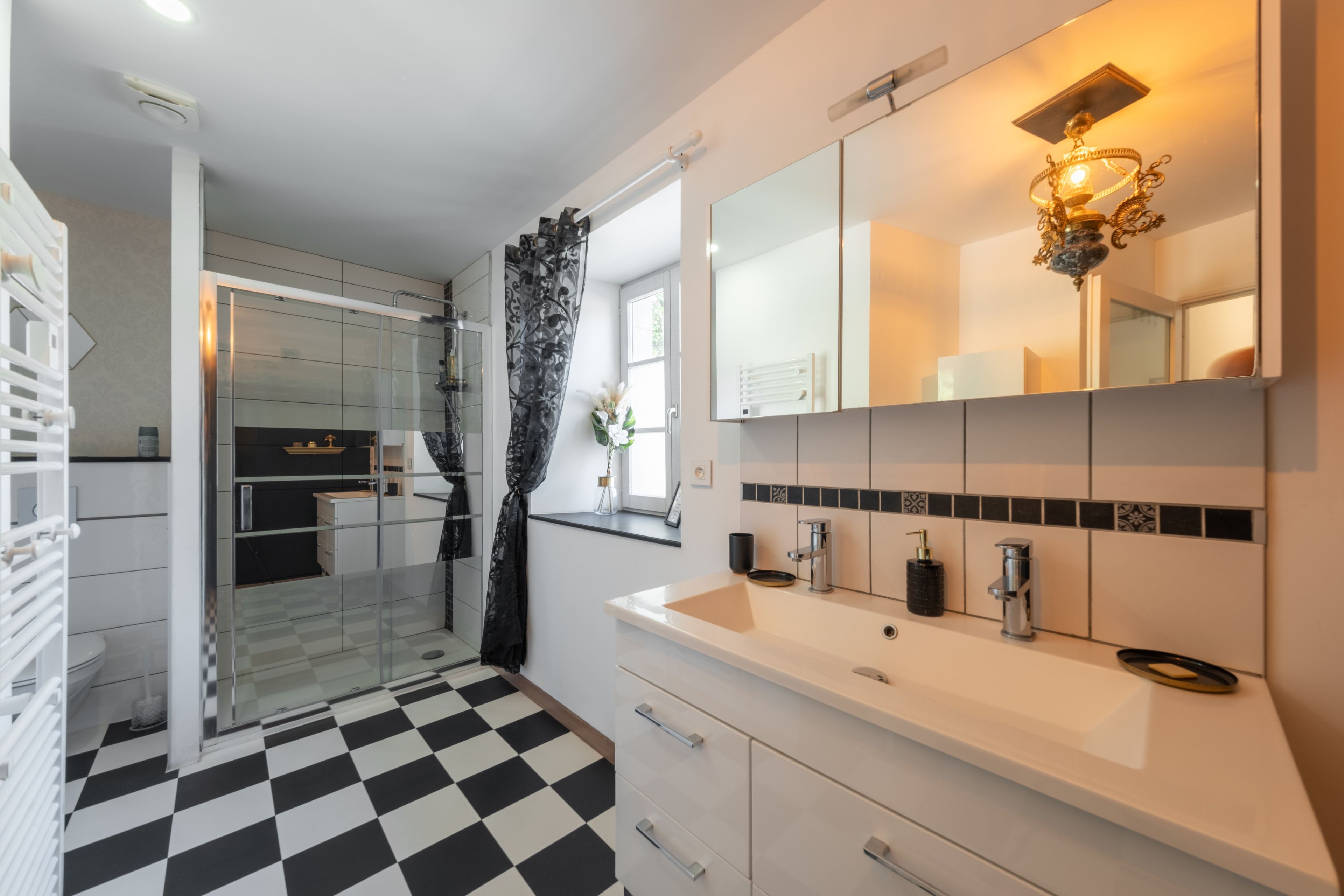 Een moderne badkamer met een zwart-wit schakeerdvloer, een glazen douchecabine en een witte wastafel met een spiegel en meerdere kranen.