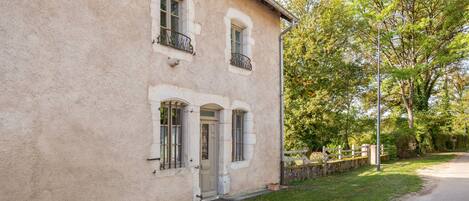 Un immeuble de deux étages avec une façade en pierre, un petit porche et une rue bordée d’arbres.