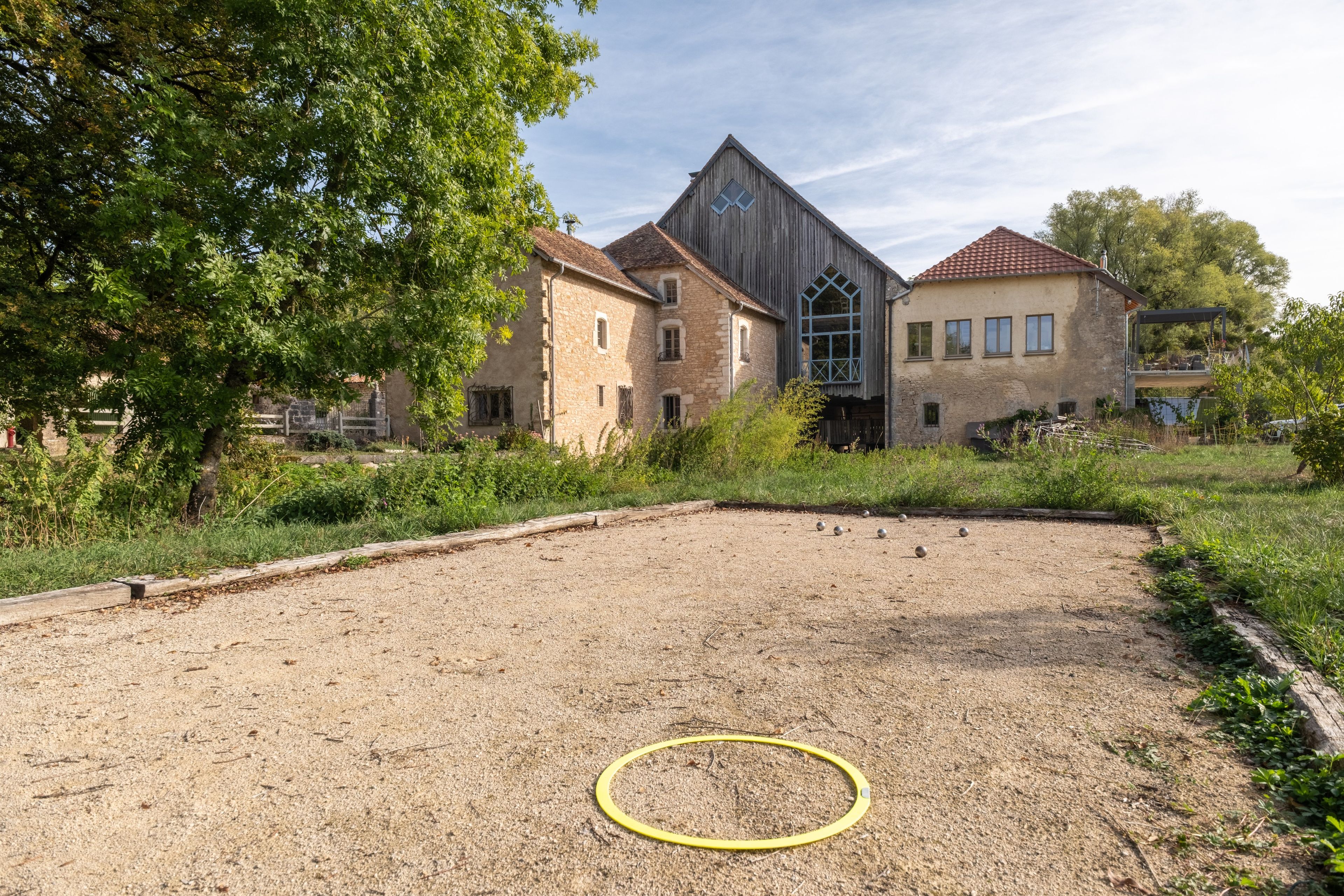 Une cour avec un sol en terre battue, un cerceau de hula-hoop jaune, et un bâtiment doté d’une façade en pierre et d’une structure en bois surmontée d’un toit triangulaire.