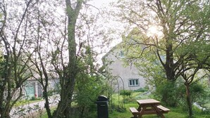 En gräsyta med ett picknickbord och en soptunna, omgiven av träd och en byggnad i bakgrunden.